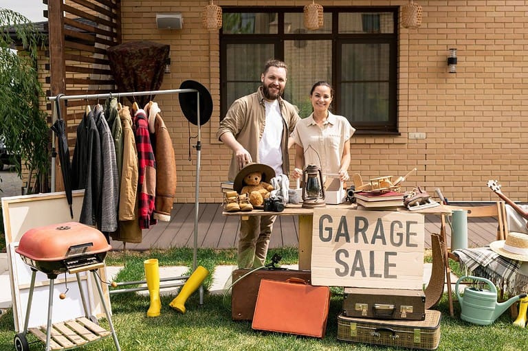 Friendly couple smiles at their garage sale. Items are available. They look happy it is a sunny day.