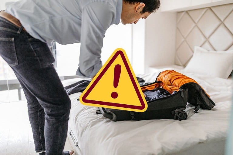 Man unpacking luggage on hotel room bed with emoji for NO. Ewwww! You'll never guess why you should put your luggage in the hotel room bathtub! The reason why will shock you!
