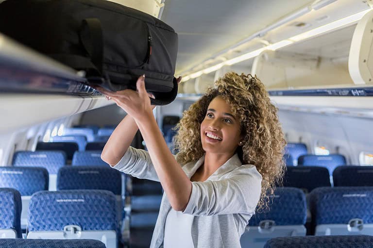 Woman putting luggage in overhead bin. Discover the insider tips that will make you an overhead bin master and keep flight attendants - and your fellow travelers - happy!