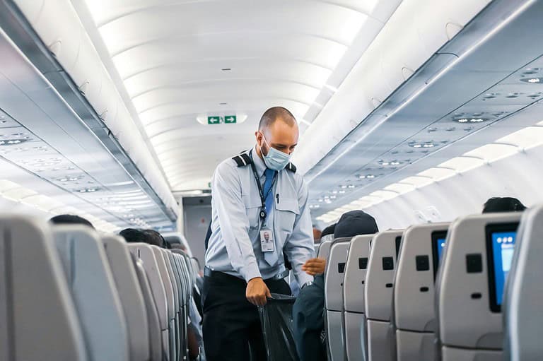 Flight attendant with mask cleaning an airplane. After hearing what a flight attendant told us, you'll never look at your airplane seat the same way again! Read this jaw-dropping exposé now!